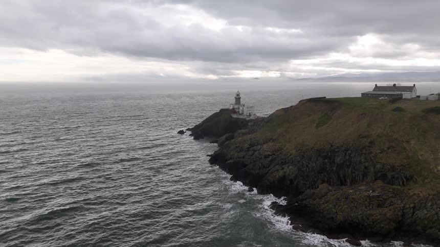 An aerial view of a lighthouse standing sentinel on a rugged cliffside as waves crash against the shore on a moody, overcast day