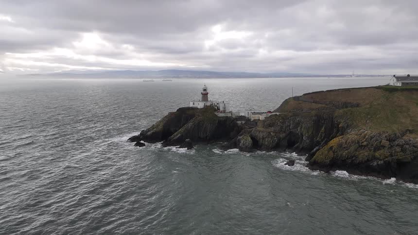 Slow aerial pullback reveals a scenic lighthouse and coastline on a moody, overcast day as waves crash against the rocks