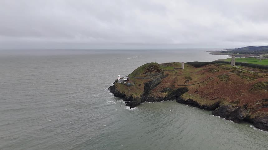 A static aerial view of the Wicklow Head Lighthouse and historic towers on a rugged Irish coastline under an overcast sky