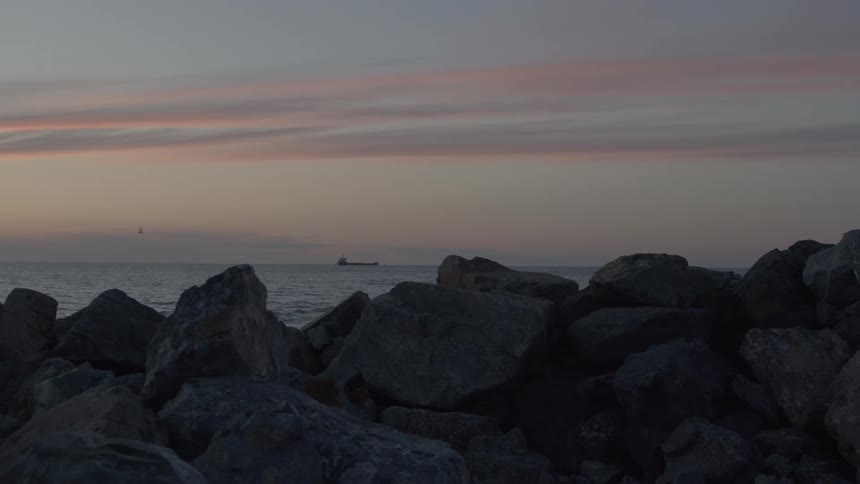 A cargo ship sits on the tranquil ocean horizon beneath a colorful twilight sky, viewed from behind a rocky jetty