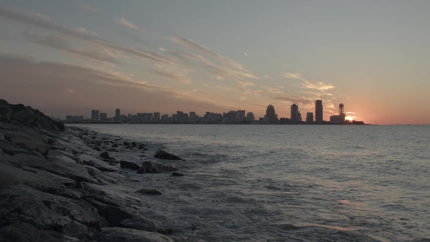 The sun sets behind a modern city skyline across the water, with waves crashing on a rocky shore in the foreground