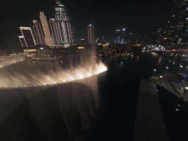 A mesmerizing aerial view captures the illuminated Dubai Fountain show dancing at night against the glittering city skyline and Burj Khalifa