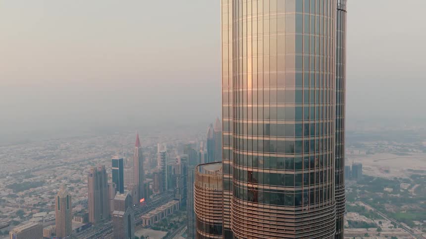 An aerial view reveals the sunset reflecting off the glass of the iconic Burj Khalifa, with the hazy Dubai skyline in the background