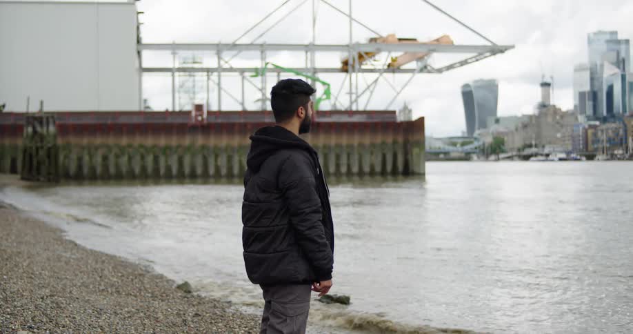 A contemplative man in a black jacket stands on a pebbly beach, looking out over the River Thames towards the London skyline on an overcast day