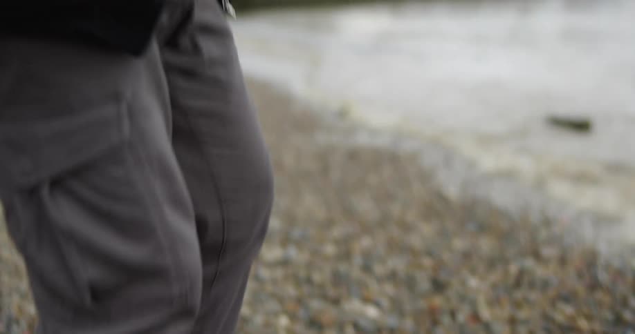 In slow motion, a person carefully selects a smooth pebble from a rocky shoreline before standing up to face the water