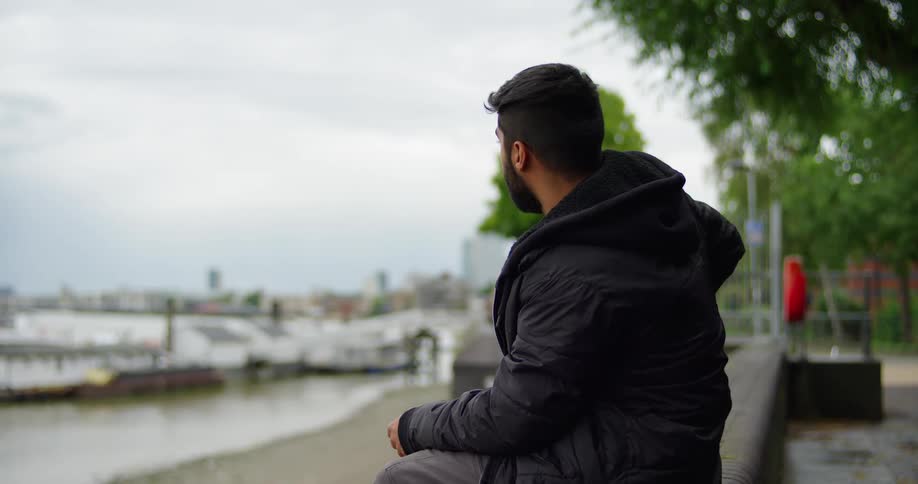A thoughtful young man in a black jacket sits by the city harbor, pausing from his phone to look out at the water on a cloudy day