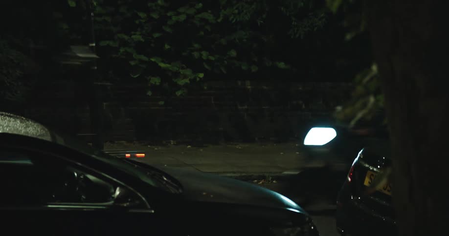 A quiet, dark street at night is briefly illuminated by the blurred lights of a passing motorcycle