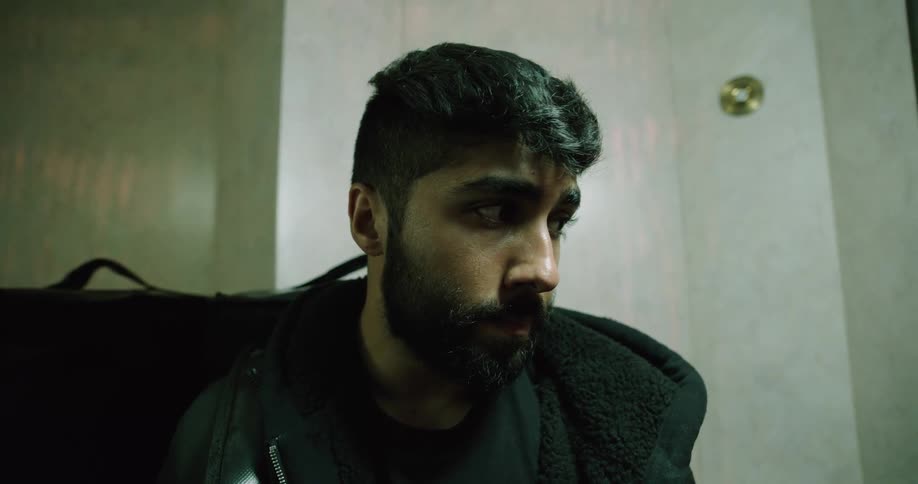 A slow-motion close-up shot of a thoughtful food delivery courier with a backpack waiting in an elevator