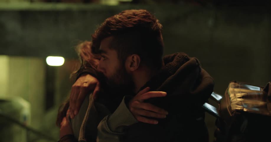 A young couple shares a tender and emotional goodbye kiss and hug in a dark city underpass at night