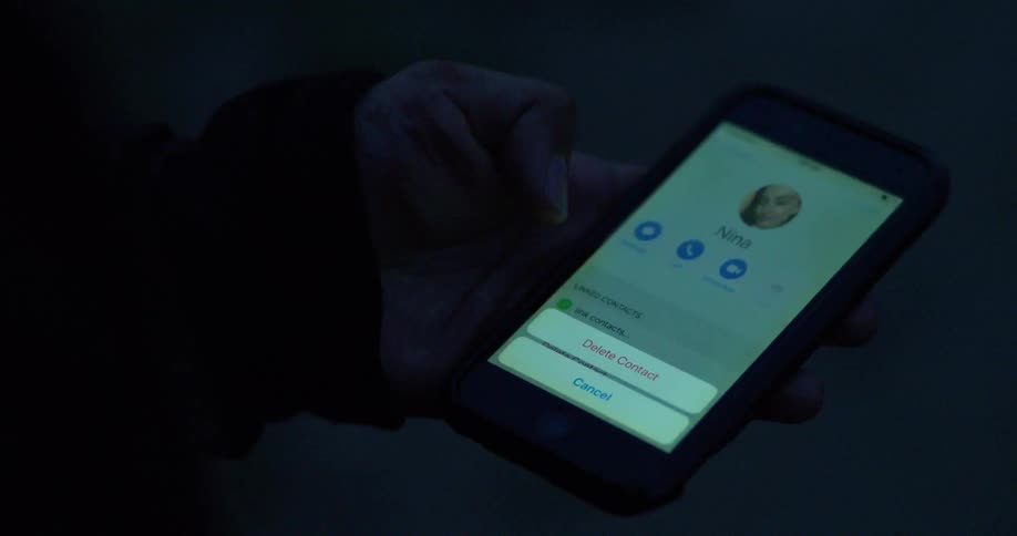 A person with a wounded hand deletes a contact from their smartphone in a dark setting, symbolizing a painful breakup