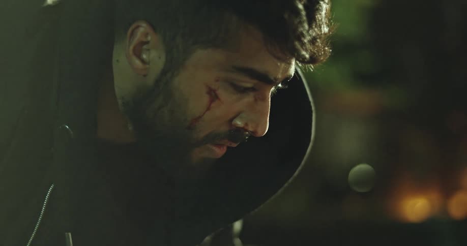 Dramatic close-up of a wounded man in a black hoodie looking down at night as the camera blurs