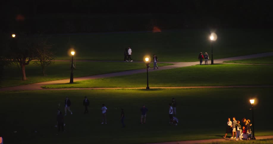 A wide shot shows various groups of people relaxing and walking through a beautifully lit park after dark