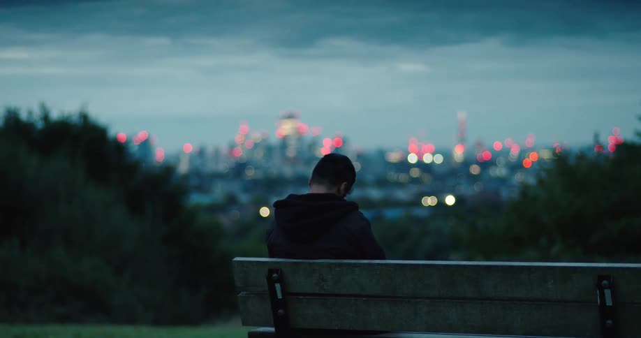 A lonely man sits on a park bench at dusk, contemplating the city skyline with out-of-focus lights in the background