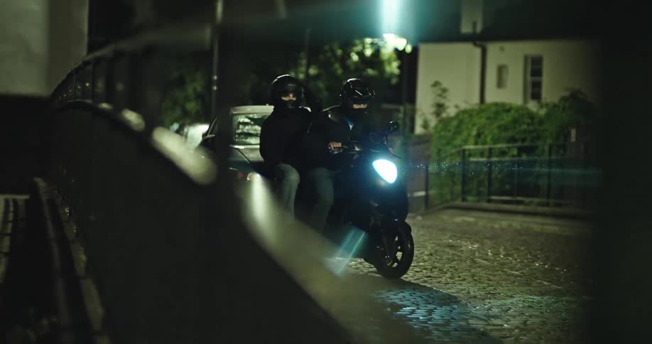 Two mysterious figures wearing helmets ride a scooter in slow motion down a dark, cobblestone street at night