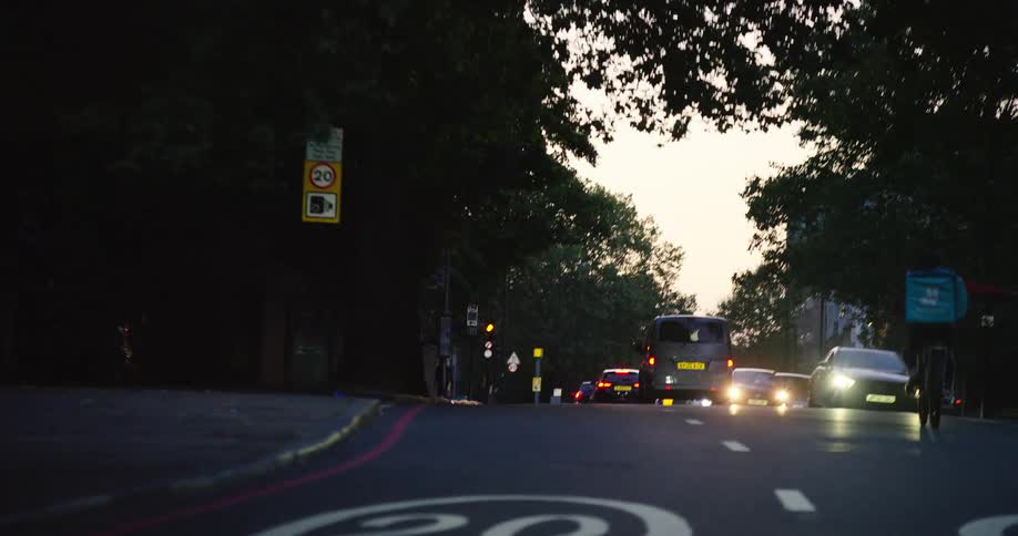 A shaky handheld shot moves down a tree-lined city street at dusk, following traffic and a delivery cyclist