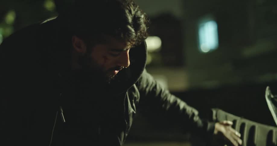 A desperate and injured young man in a dark hoodie searches through a food delivery bag in a dim city alley at night