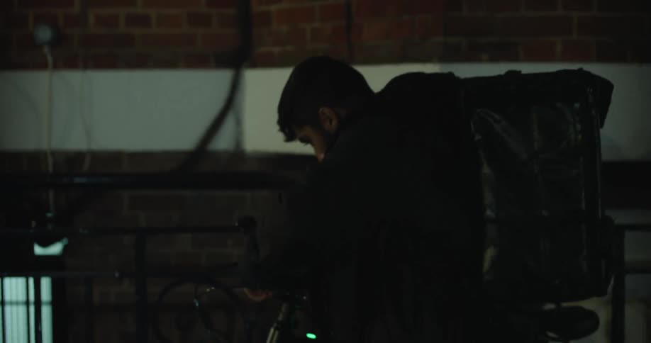 A young male food delivery driver leaves a building at night, checks his smartphone, and rides away on his bicycle