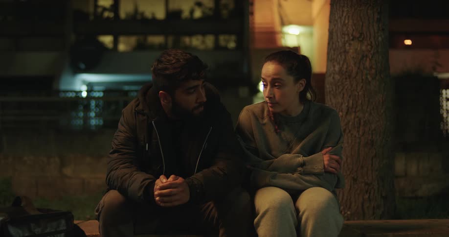 A male and female food delivery driver have a serious talk while sitting on a curb at night before getting up to leave