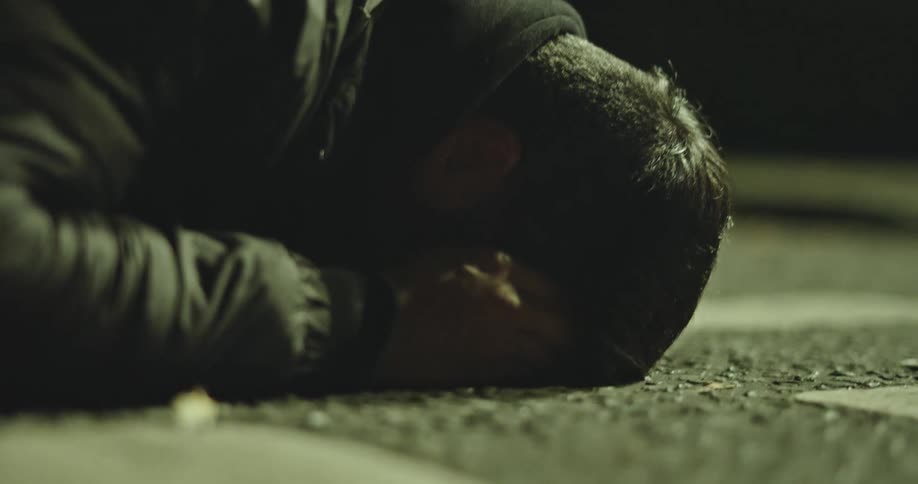 A dramatic close-up of a distraught man lying face down on the pavement at night