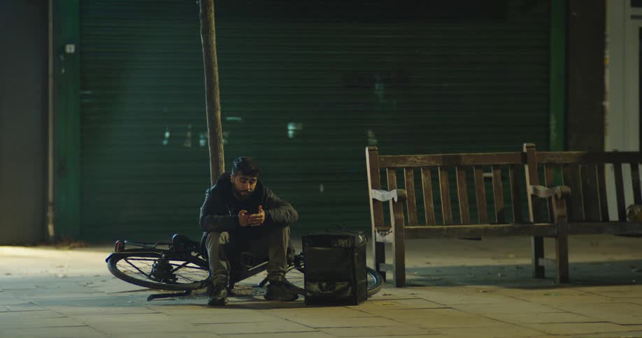 A male food delivery cyclist sits on the pavement next to his bicycle and delivery bag, taking a break on an empty street at night