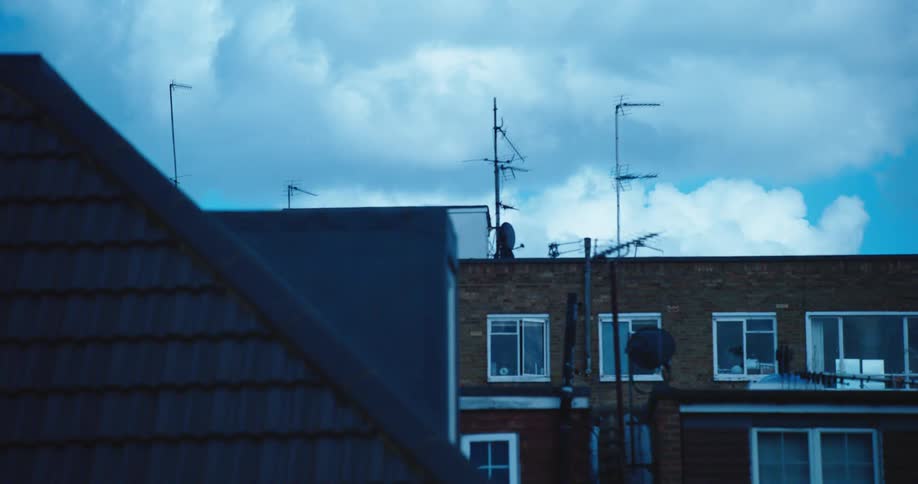 A static shot of quiet suburban rooftops with television antennas under a dramatic, cool-toned sky