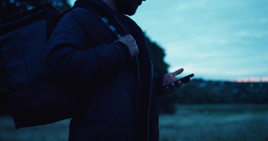 An injured man with a duffel bag walks through a field at dusk and checks his smartphone in slow motion