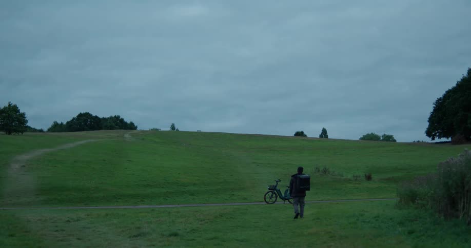 A delivery driver with a large backpack walks to his electric bike in a wide-open park on an overcast day