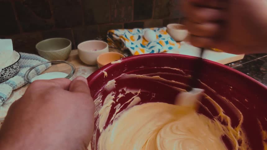 A first-person view of hands vigorously mixing creamy cake batter in a red bowl, with baking pans ready on the stove in the background