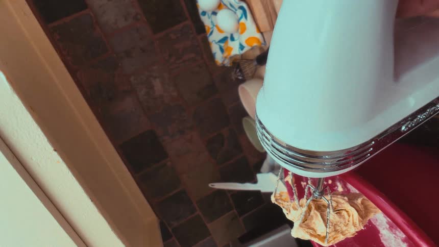 An overhead vertical view of an electric mixer blending batter in a red bowl, with baking ingredients laid out on the counter