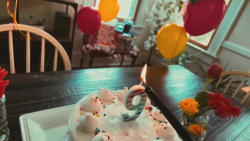 A lighter ignites the number nine candle on a white birthday cake in slow motion, surrounded by colorful balloons and flowers in a cozy dining room