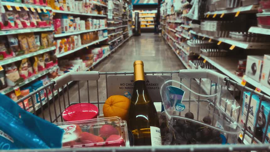 A point-of-view shot from a shopping cart filled with wine and fruit as it moves down a long grocery store aisle