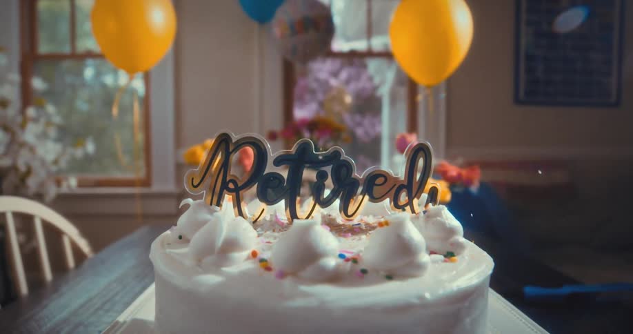 A festive retirement cake with a 'Retired' topper sits on a table during a celebration with balloons in the background