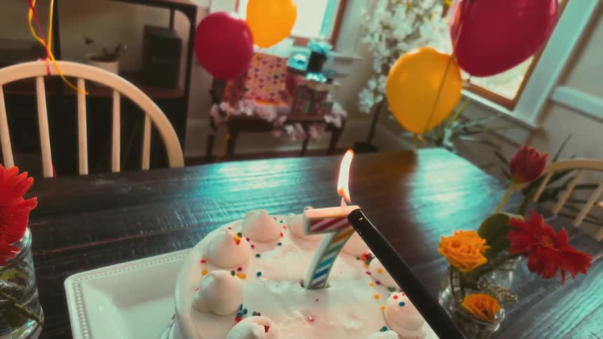 A first-person view of a long lighter igniting the number seven candle on a white frosted birthday cake at a party