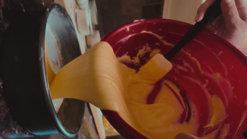 A slow-motion, top-down shot shows thick, yellow cake batter being poured from a mixing bowl into a baking pan on a messy kitchen counter