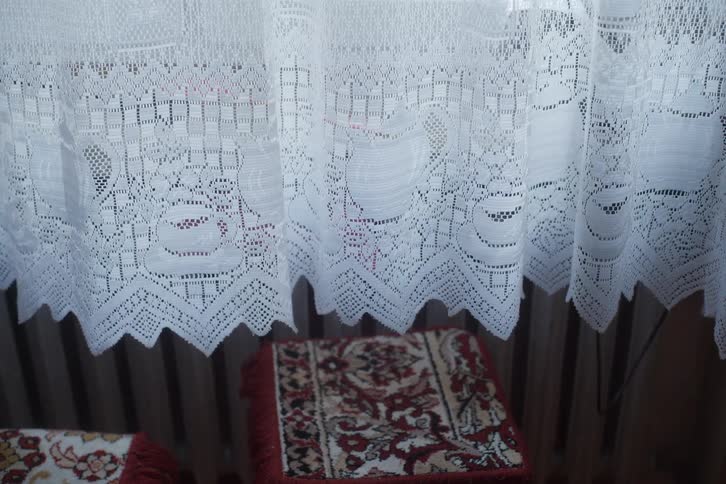 A delicate white lace curtain with an intricate teapot pattern sways gently in a vintage-style room