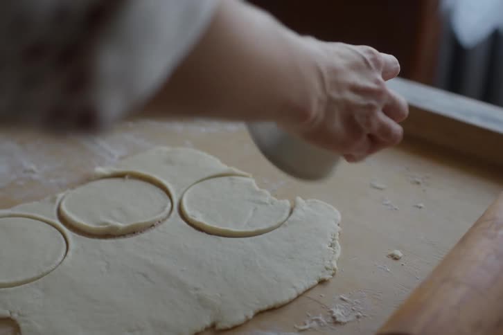 Close up of a baker's hands using a round cutter to shape circles out of rolled dough on a floured wooden surface