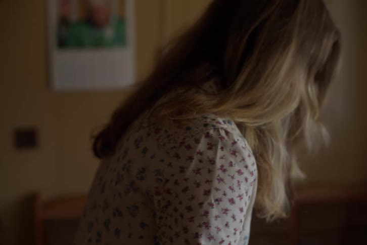 Close-up over-the-shoulder shot of a woman with long blonde hair in a floral shirt, seen from behind in a dimly lit room