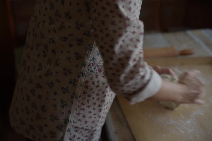 A woman in a floral apron kneads dough by hand on a wooden surface dusted with flour