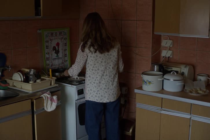 A woman with long hair lights a gas stove to cook a meal in a dimly lit, modest kitchen
