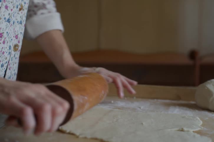A baker's hands meticulously roll out fresh dough with a wooden rolling pin on a floured surface in slow motion
