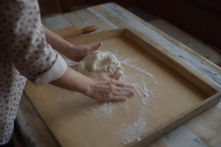 A woman spreads flour on a wooden board before expertly kneading a ball of fresh dough by hand