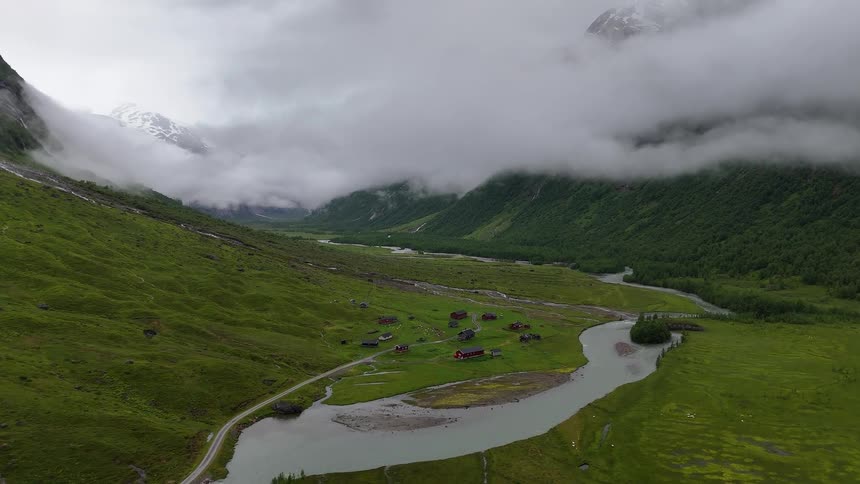 A static aerial view shows a picturesque cluster of red cabins in a foggy, green mountain valley next to a winding river in Norway