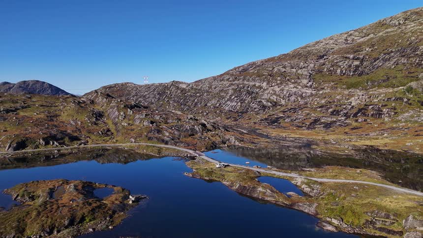 A static aerial view shows a camper van parked on a causeway crossing a tranquil blue lake in a remote, rocky mountain landscape