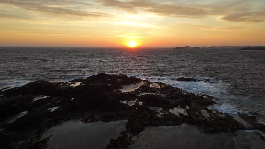 Aerial pullback shot revealing a rugged rocky coastline as waves crash against the shore during a beautiful golden sunset over the ocean