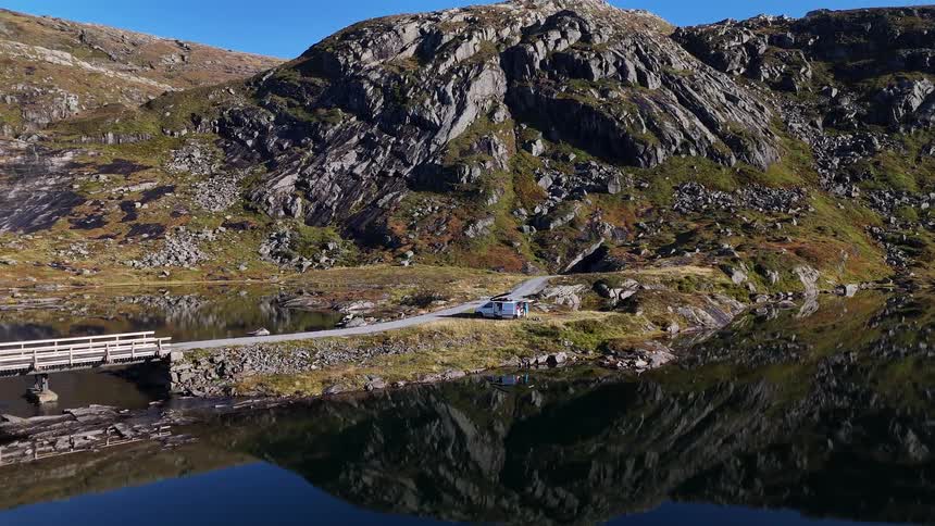 Aerial footage captures a serene scene of a camper van parked on a remote mountain road beside a tranquil, reflective lake in Norway