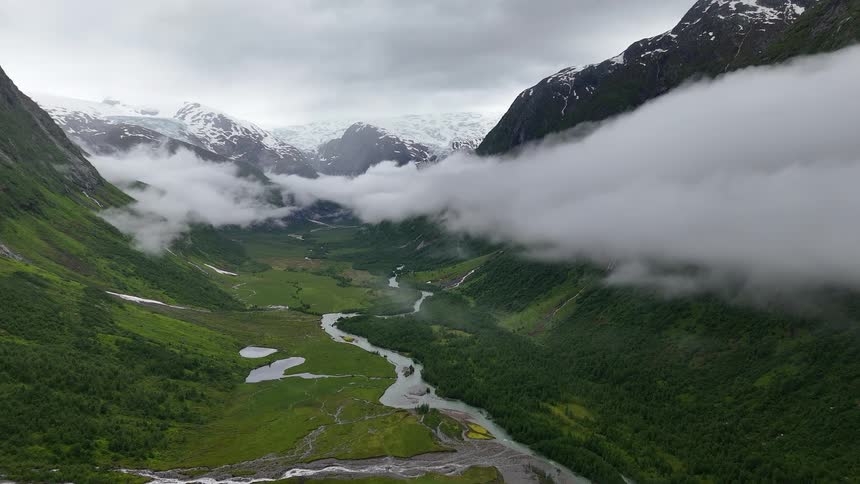 An epic aerial view of a lush green mountain valley with a winding river leading to a distant glacier under a cloudy sky