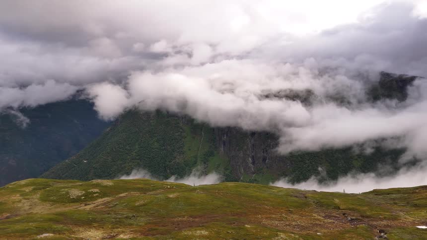 From a high tundra plateau, a majestic mountain range is enveloped in a thick blanket of rolling clouds and mist