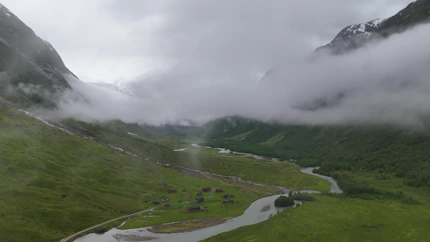 A dramatic aerial view shows a secluded village in a lush green Norwegian valley, with a winding river and thick clouds drifting below majestic mountains