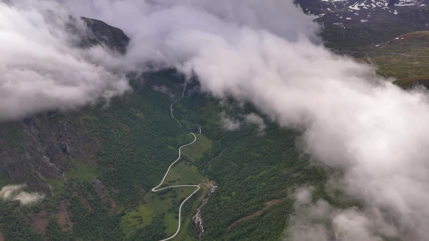 Flying above the clouds over a dramatic green mountain valley with a winding road and river below
