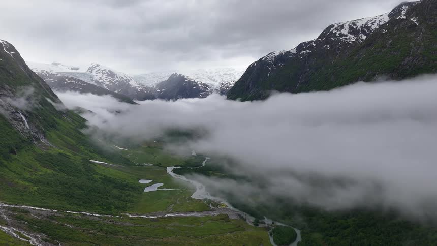 A slow aerial pullback reveals a stunning green valley and river below the clouds, with snow-capped mountains and a glacier in the distance